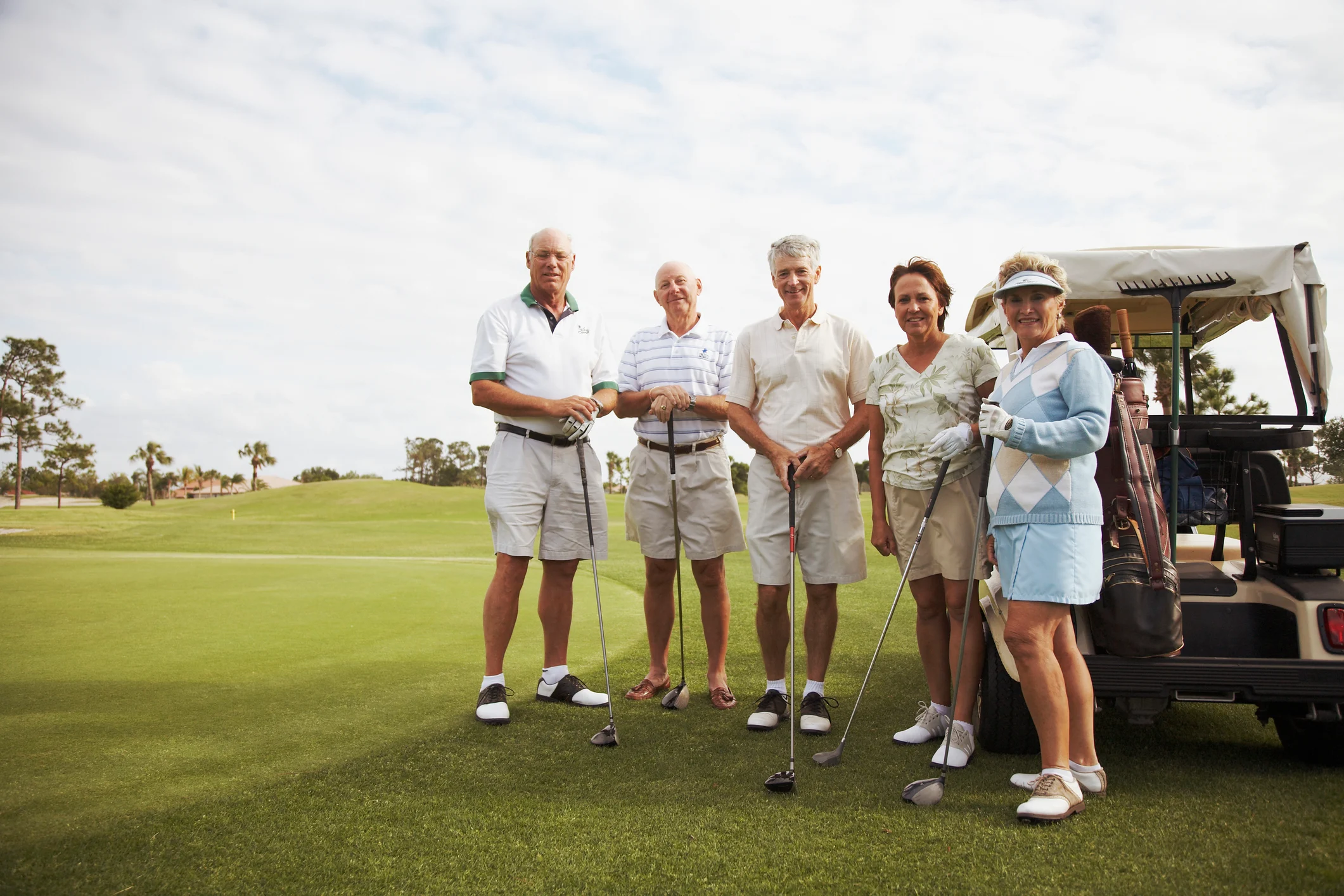 A group of five older adults in golf attire standing on a golf course with tennis golf clubs and a golf cart in the background.