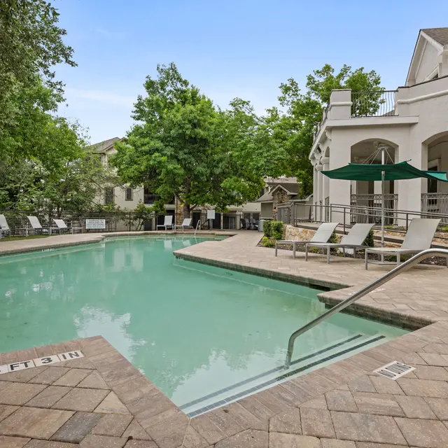 A serene swimming pool area surrounded by trees, with lounge chairs and an umbrella. The pool has steps leading in, and the setting appears calm and inviting.