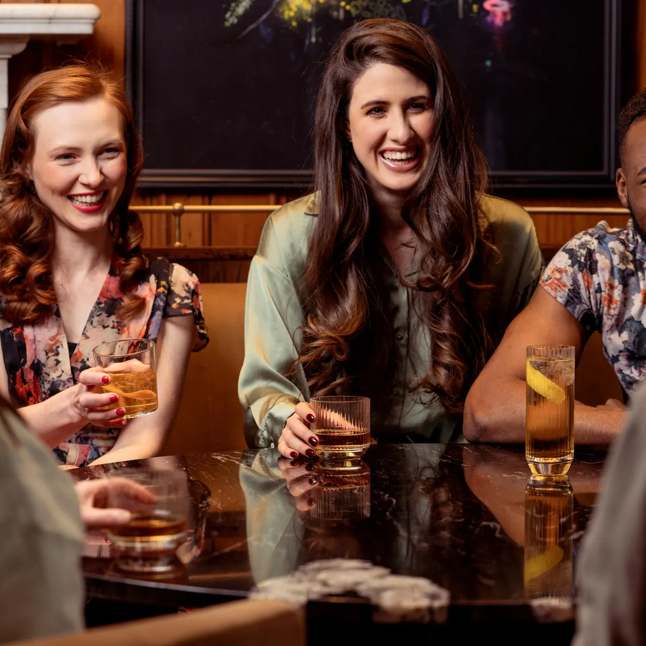 A group of friends laughing and enjoying drinks around a table in a cozy setting.