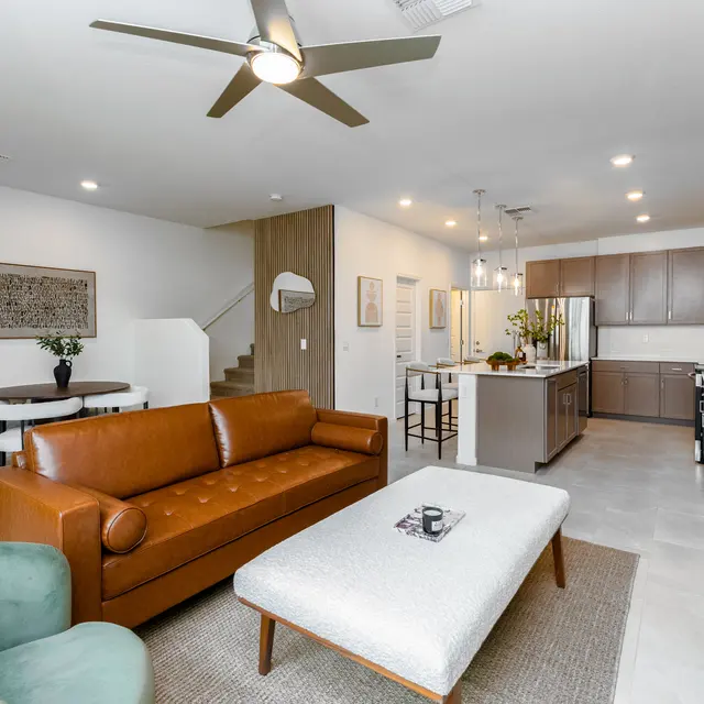 A modern living room featuring a brown leather sofa, a light-colored coffee table, and green armchairs. The space includes a dining area and a kitchen with wooden cabinets. A staircase is visible in the background along with decorative wall art.
