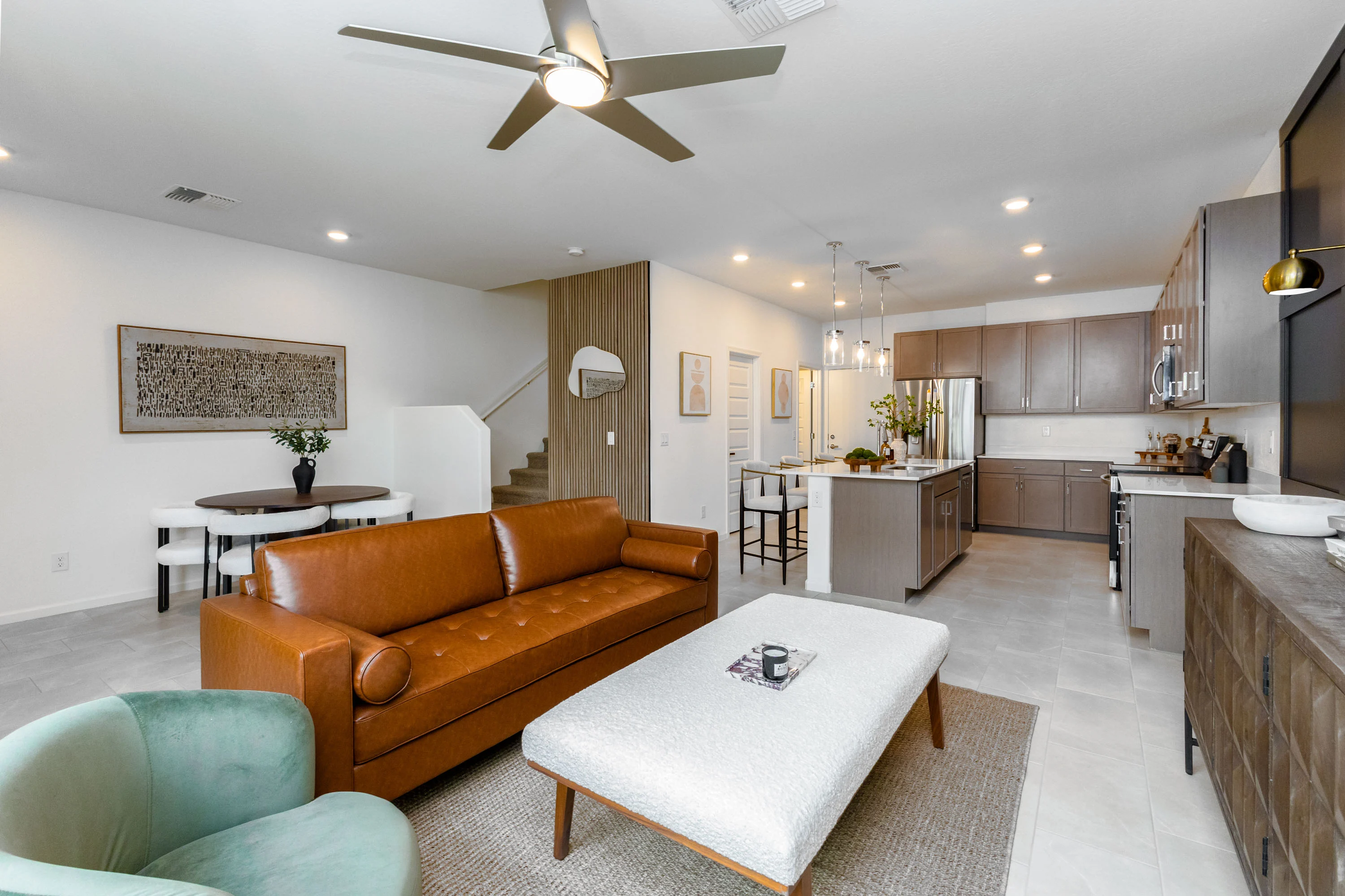 A modern living room featuring a brown leather sofa, a light-colored coffee table, and green armchairs. The space includes a dining area and a kitchen with wooden cabinets. A staircase is visible in the background along with decorative wall art.