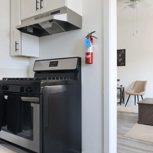 A modern kitchen featuring a black gas stove with an oven, a range hood above it, and a fire extinguisher mounted on the wall. In the background, there is a cozy living area with a beige chair and a coffee table.