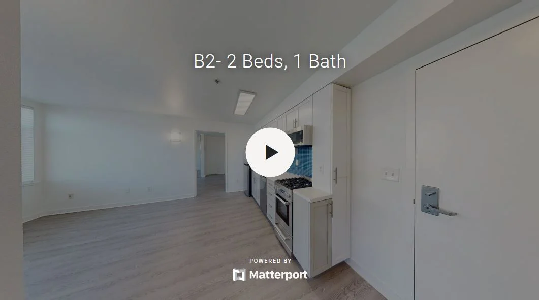 A virtual tour image of a modern apartment featuring a spacious layout with a kitchen area visible on the right and a living space extending towards the left.