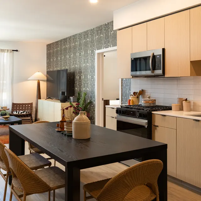 A modern apartment featuring an open layout with a kitchen and dining area. The kitchen has light wooden cabinets and a black dining table with chairs. There is a sofa and a lamp in the living area, along with decorative plants and a television.