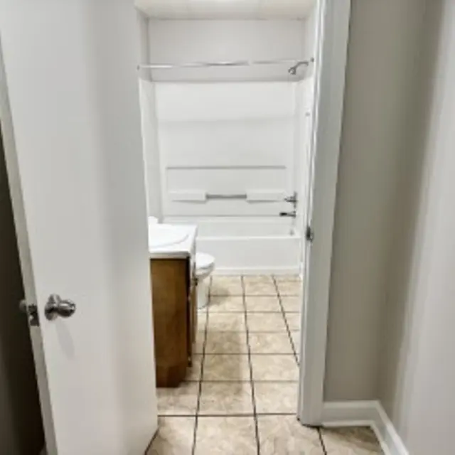 View of a bathroom from a hallway, featuring a bathtub, a toilet, and a sink with a wooden cabinet. The flooring is tiled and the walls are painted light colors.