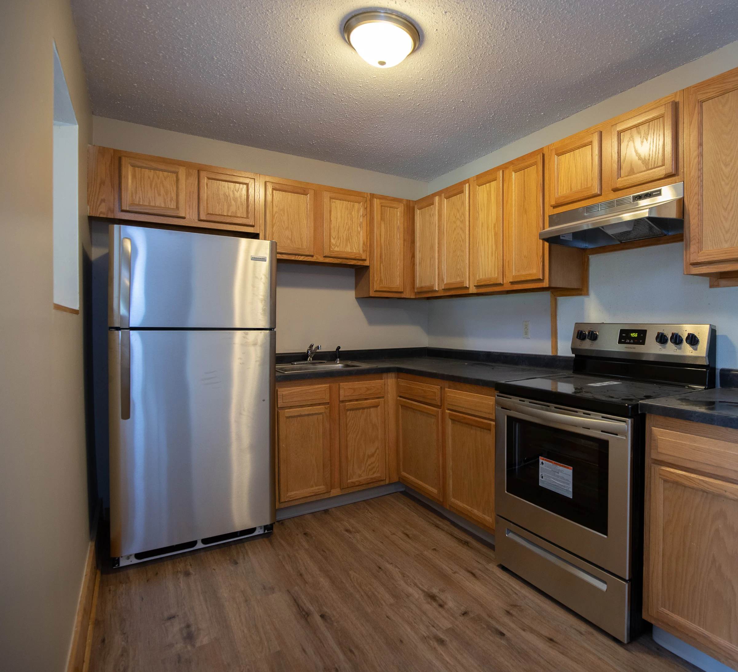 A modern kitchen featuring wooden cabinetry, stainless steel appliances, a black countertop, and a warm-toned floor.