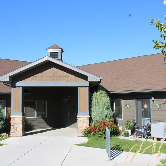 A residential building entrance with a paved walkway leading to the front door. Flanked by green shrubs and decorative flowers on either side, the building features a brown wooden exterior and a gray roof under a clear blue sky.