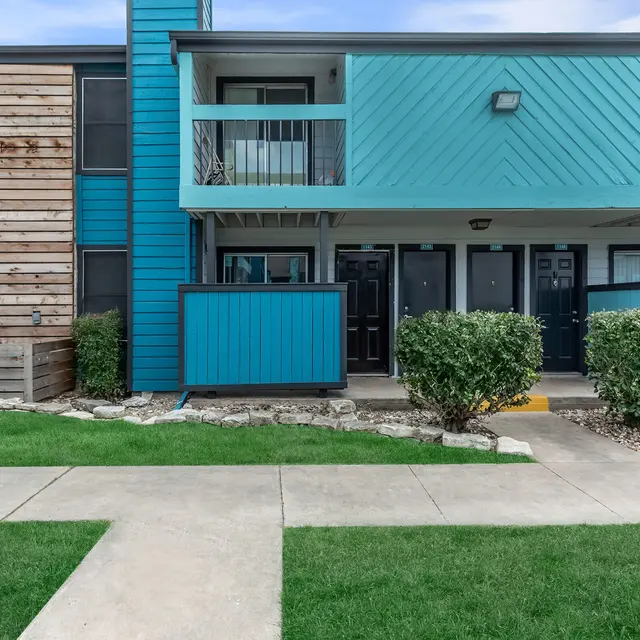 A modern apartment complex with vibrant blue siding and wooden paneling, featuring a well-maintained lawn and pathway in front.