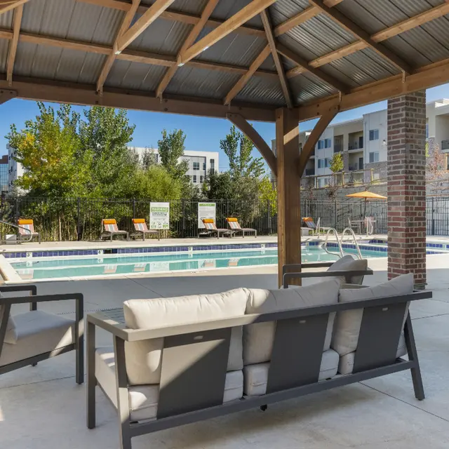 Patio Area by the Pool A comfortable patio area with gray outdoor sofas under a wooden gazebo, overlooking a swimming pool surrounded by green trees and lounge chairs.