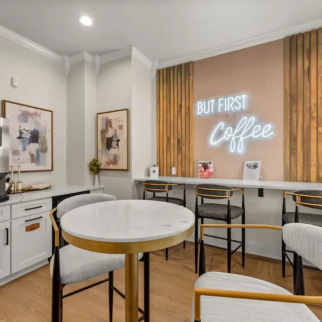 A modern coffee corner featuring a brewing machine, a round marble table, and stylish chairs. The wall displays a neon sign that reads 'BUT FIRST Coffee'.