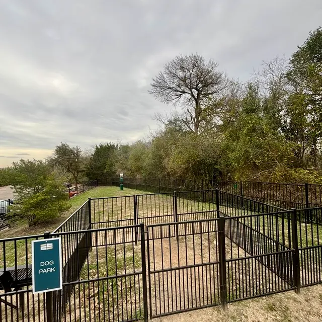 A fenced dog park with some grassy areas and a few trees in the background, under a cloudy sky.