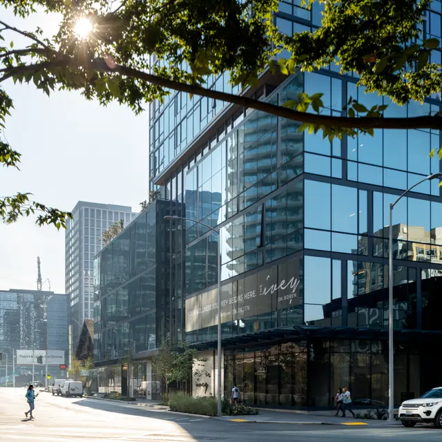 A street view of a modern, glass-fronted building with reflections of nearby skyscrapers and trees in the foreground. The scene is bright with sunlight, showing a clear blue sky.
