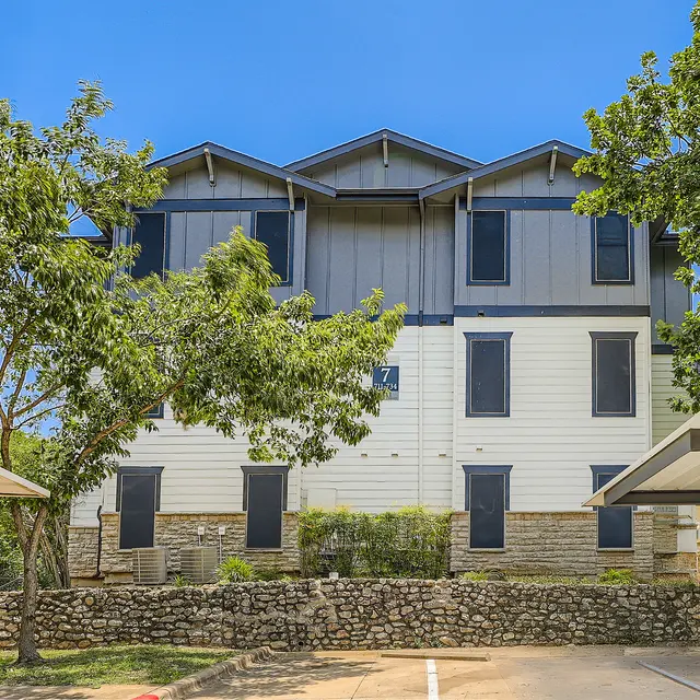 A three-story apartment building with a white exterior and blue accents, surrounded by green trees and shrubs. The building features several windows and is set against a clear blue sky.