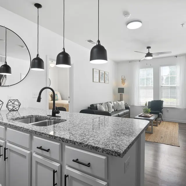 A modern apartment showing an open kitchen with a stone countertop and living area in the background. The kitchen features a sink with a stylish faucet, geometric decorations, and pendant lights. The living area has a couch, a coffee table, and a TV, with large windows letting in natural light.