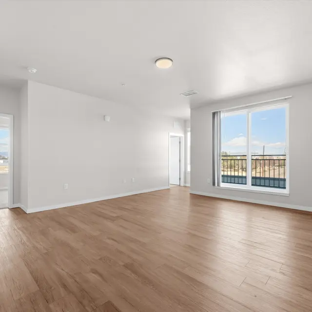 A bright and spacious room with wood flooring, large windows, and a door leading outside. The walls are painted white, and there is a ceiling light fixture.