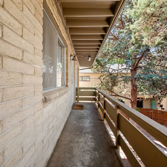 A narrow balcony walkway with a light-colored brick wall on one side and wooden railing on the other, surrounded by trees in a residential setting.