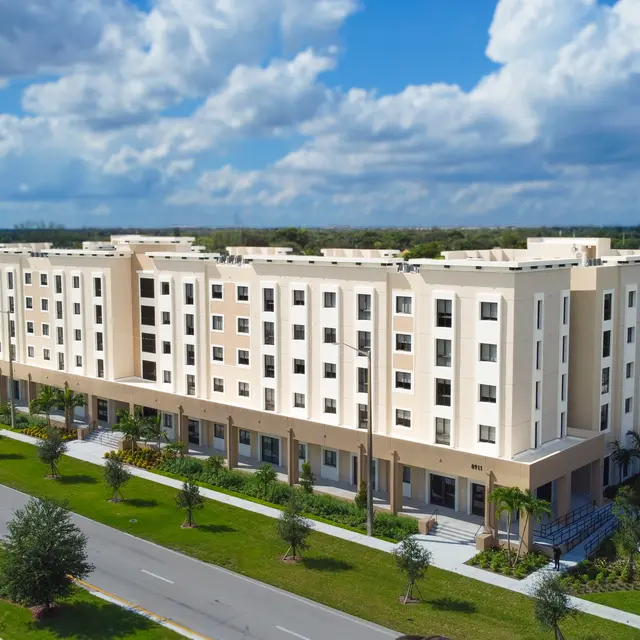 A modern multi-story apartment building with a light-colored facade, surrounded by green landscaping and palm trees, under a blue sky with fluffy clouds.