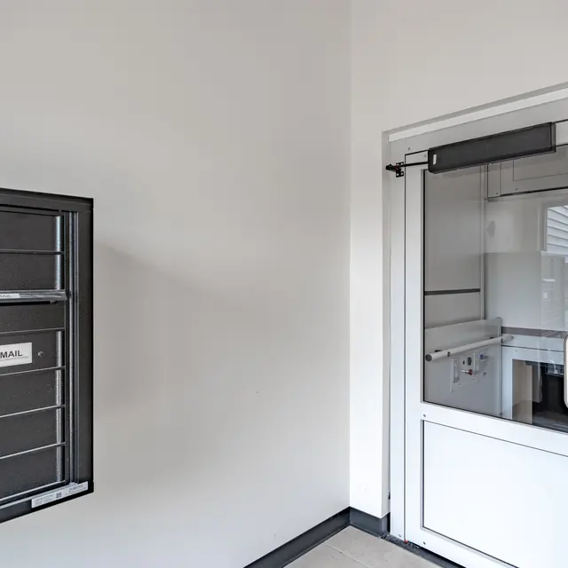 Interior view of a mail area featuring a black wall-mounted mail box with several compartments, and a glass door leading to another space.