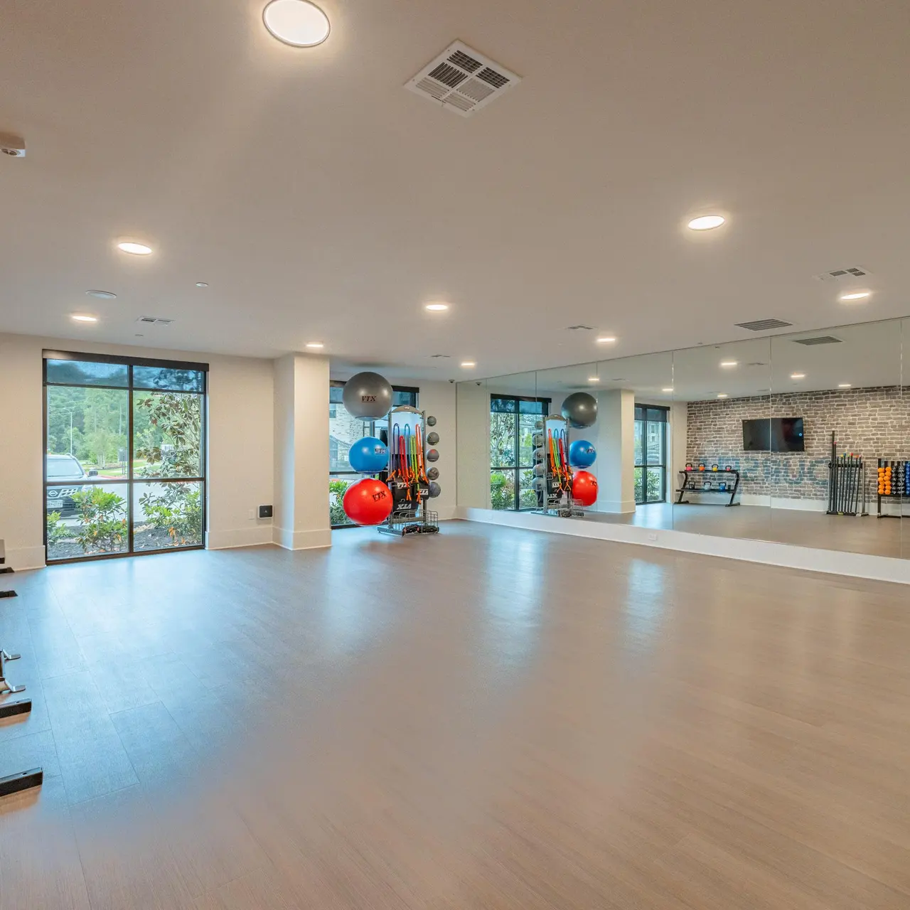 A spacious modern fitness studio featuring large windows, wooden flooring, and a mirrored wall. Various fitness equipment, including exercise balls and resistance bands, are neatly organized against the walls.