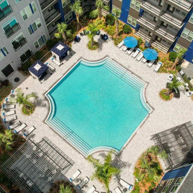 Aerial view of a pool area surrounded by modern apartment buildings, featuring palm trees, lounge chairs, and umbrellas.