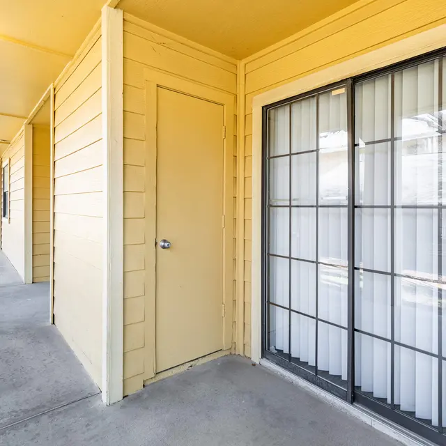A hallway in an apartment complex, featuring a closed door with a window next to it and a concrete walkway.