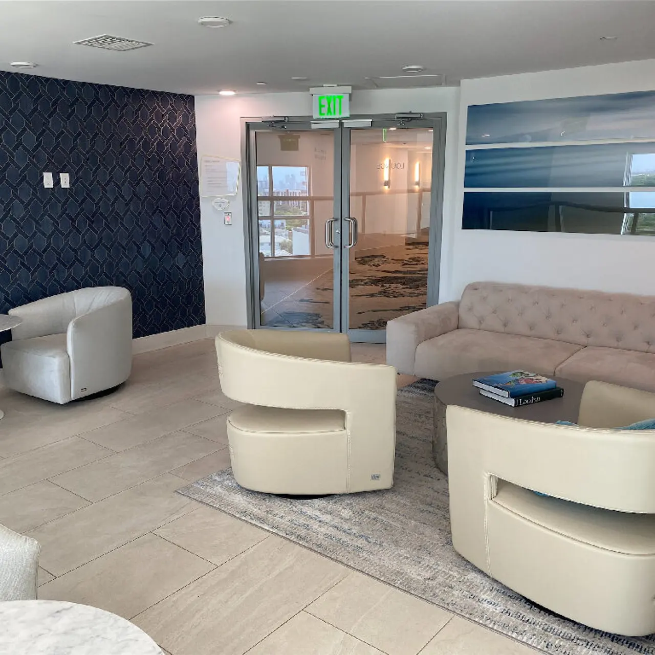 A modern lounge area featuring beige furniture, a round table, and a textured navy blue wall. There are large windows with a view outside, and a rug on the floor.