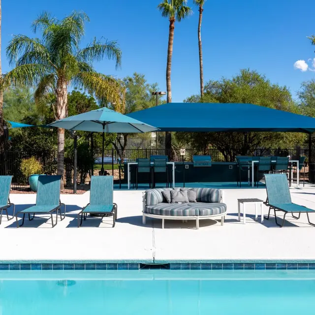 A sunny pool area featuring several lounge chairs, an umbrella, and a circular seating arrangement next to the water.