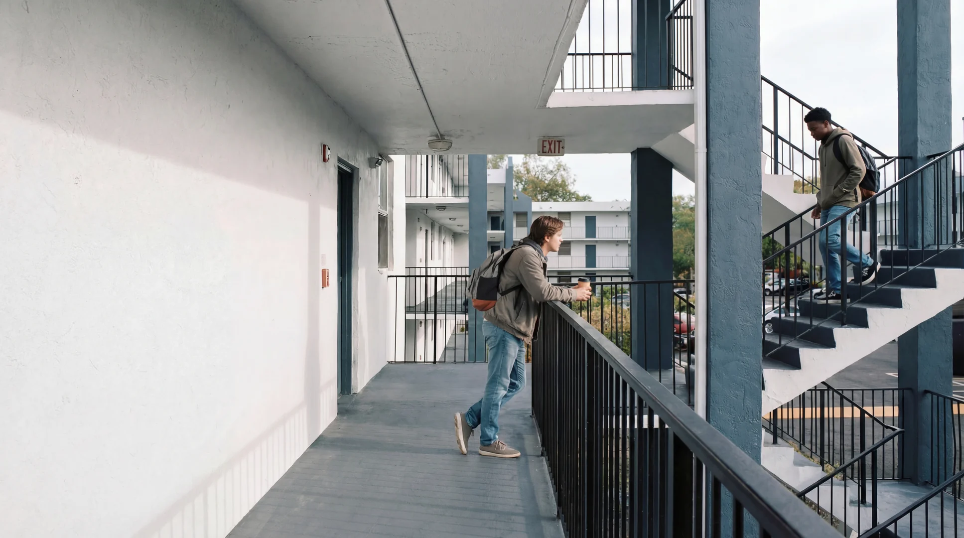 Two people interacting on a balcony outside an apartment building, with one leaning on the railing and the other walking down the stairs.