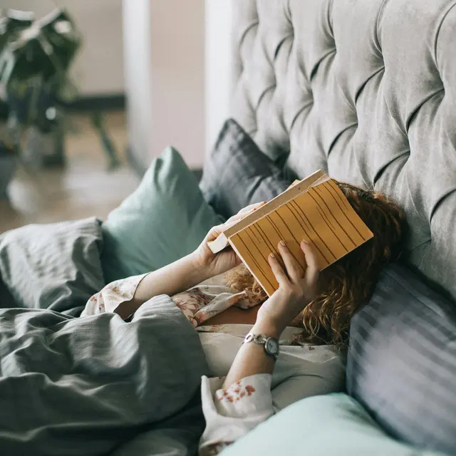 Cozy Reading in Bed A person lying in bed covered with blankets, holding a book close to their face. The bed has soft pillows in varying shades of green and gray. The background suggests a warm, cozy room with natural light coming in.
