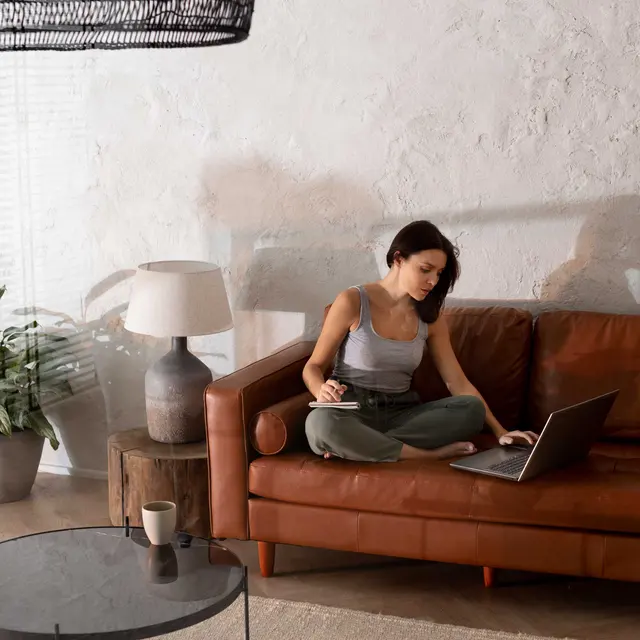 A woman sitting cross-legged on a brown leather sofa, working on a laptop, with a coffee mug beside her on a glass table.