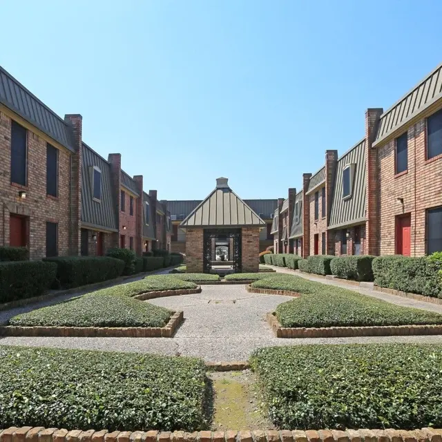 A view of a well-maintained courtyard in an apartment complex, featuring brick buildings and manicured hedges under a clear blue sky.