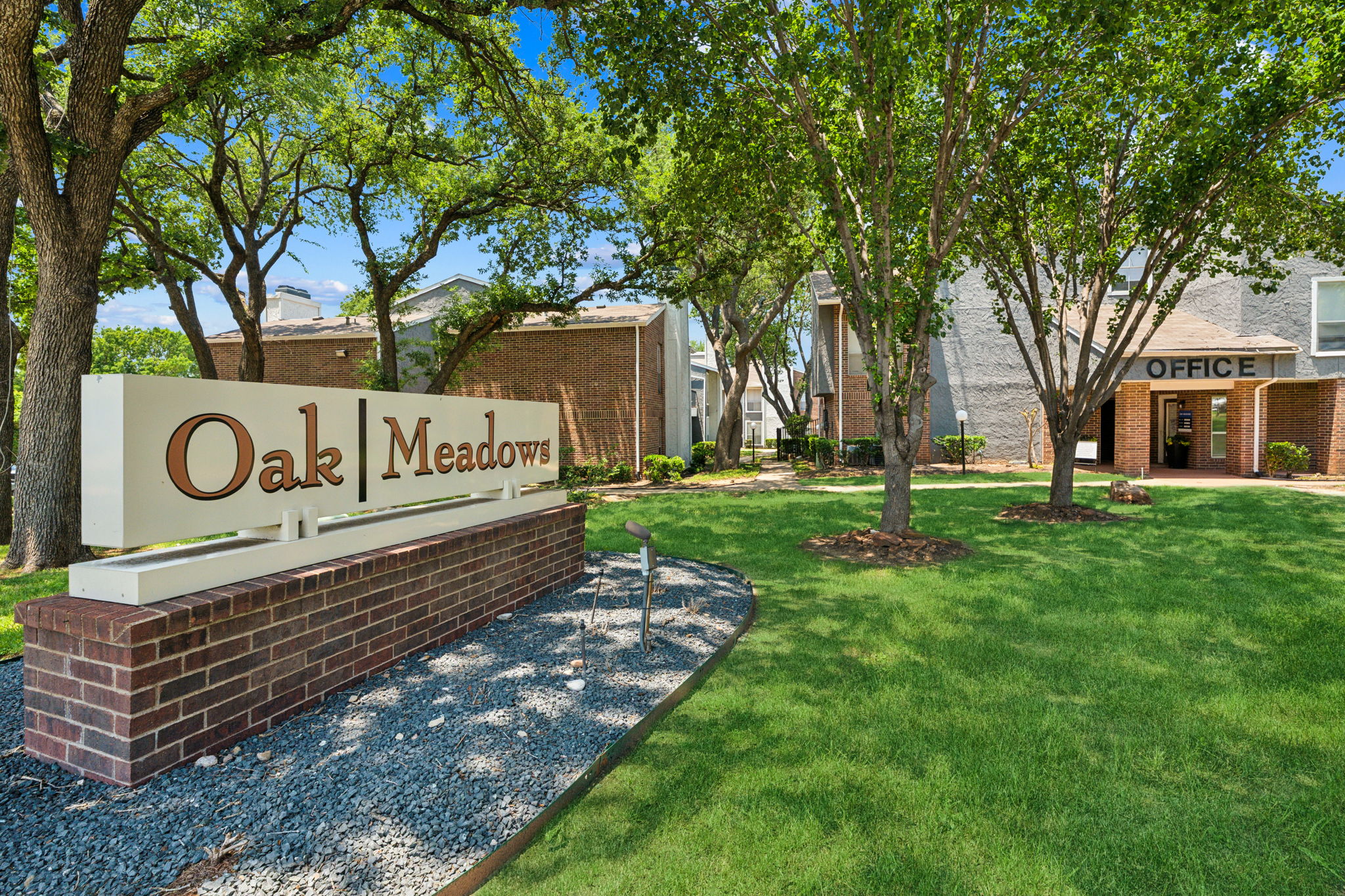 A welcoming entrance sign for Oak Meadows set in a green landscape with trees and well-maintained grounds. The office building is visible in the background.