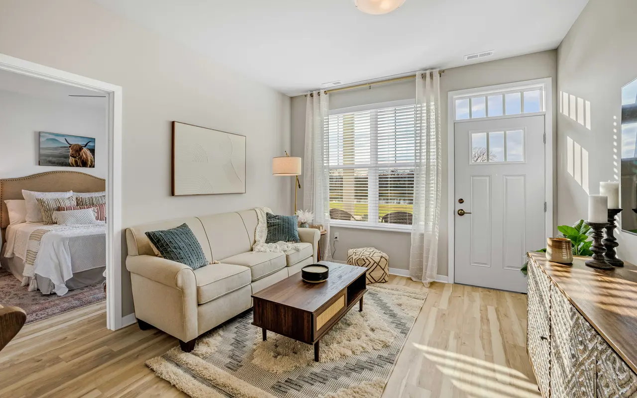 A cozy living room featuring a light-colored sofa, a wooden coffee table, and a large window with curtains allowing natural light. A bedroom is partially visible in the background.