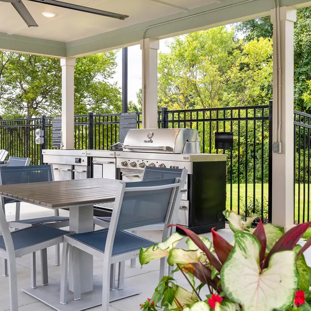 A covered outdoor patio featuring a dining table with chairs, a barbecue grill, and surrounding greenery.