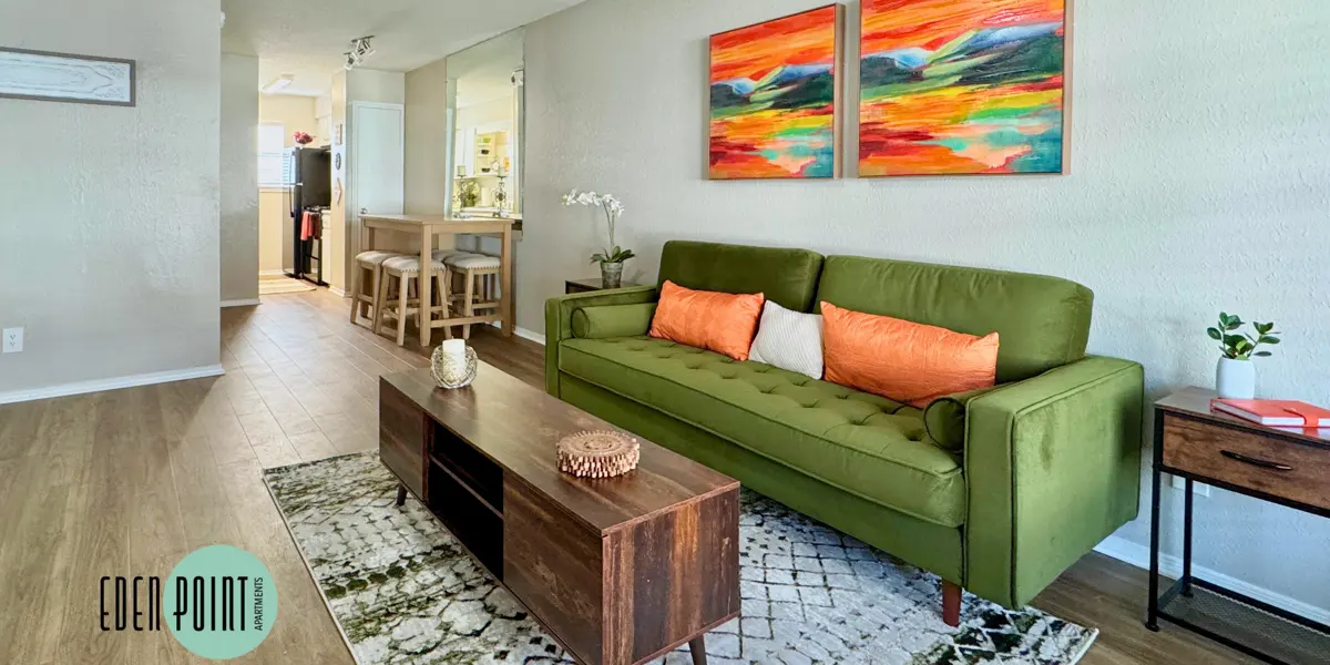 A bright, modern living room featuring a green sofa with orange pillows, a wooden coffee table, and colorful abstract artwork on the walls. The room has wooden flooring and a stylish rug underneath the coffee table.