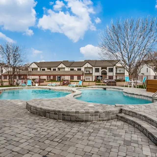 A serene pool area with two pools surrounded by lounge chairs and a view of apartments in the background under a blue sky with fluffy clouds.