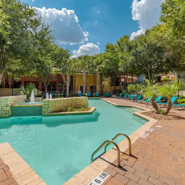 A serene swimming pool area surrounded by trees and lounge chairs, featuring a small fountain in the water and a clear blue sky with clouds.