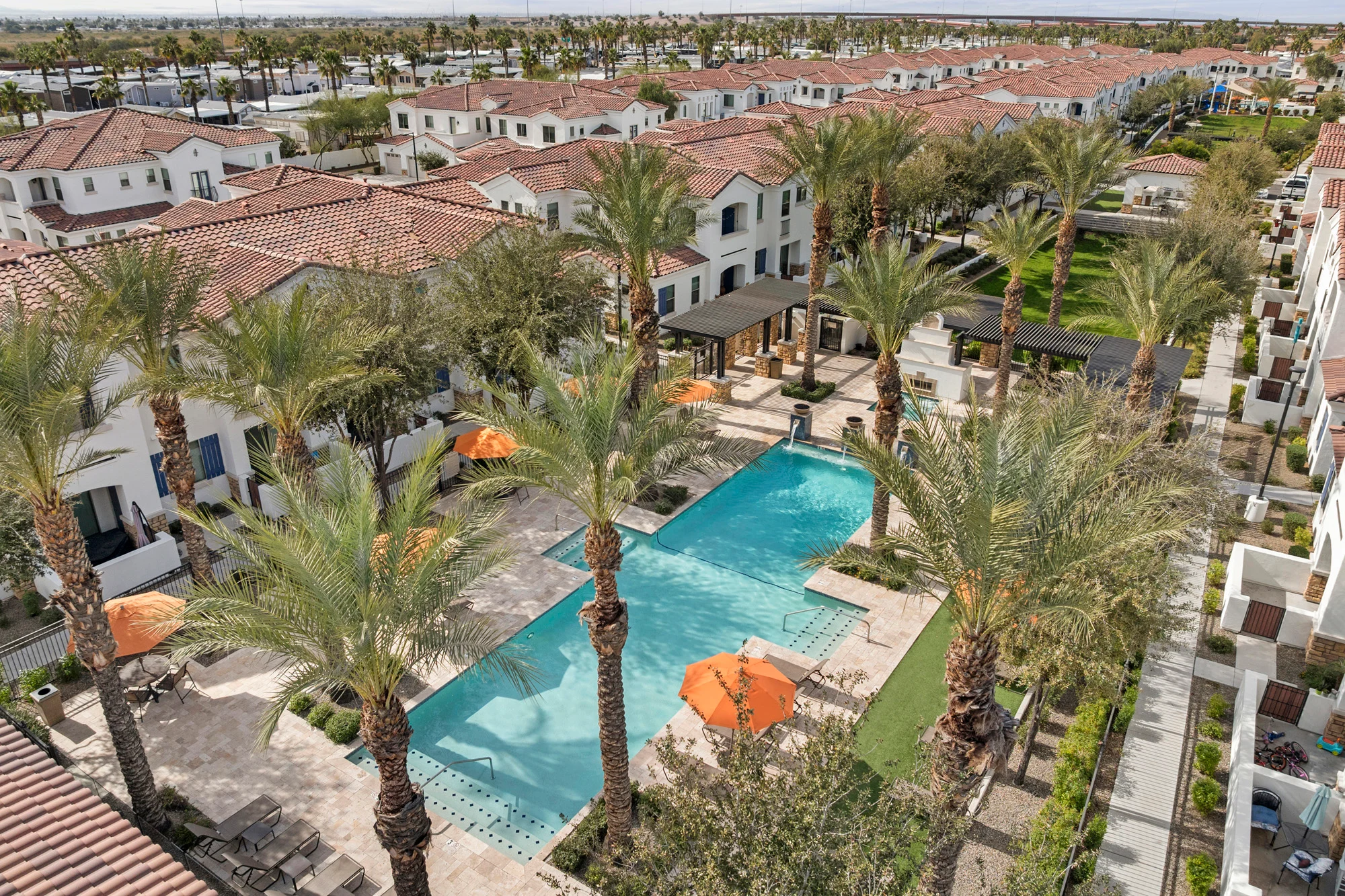 Aerial view of a resort pool area surrounded by palm trees, featuring a blue swimming pool, lounge chairs, and orange umbrellas, with residential buildings in the background.
