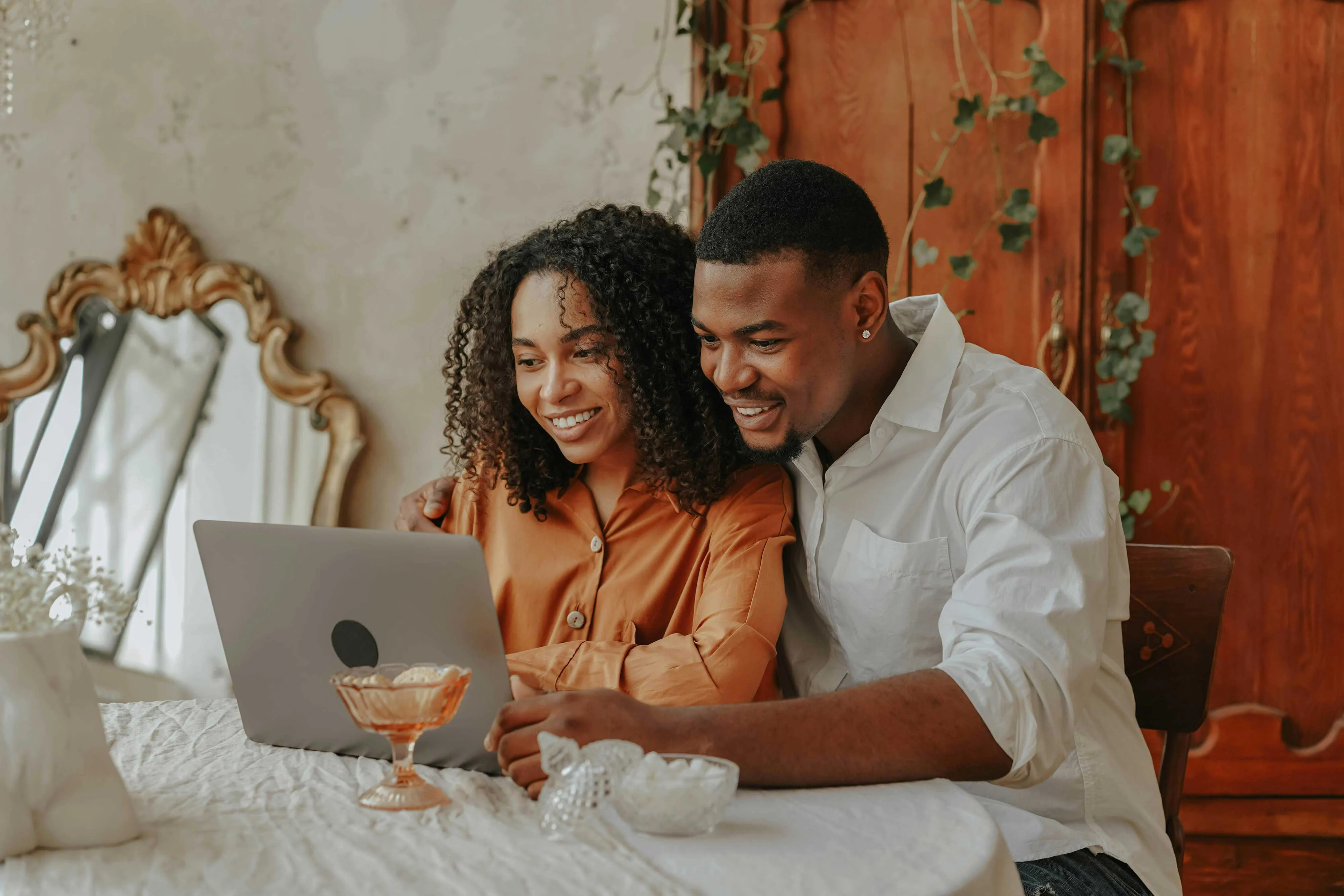 A couple sitting at a table, smiling and looking at a laptop together in a cozy indoor setting. The woman has curly hair and is wearing a peach-colored shirt, while the man, with a short hairstyle, is in a white shirt. They appear engaged and happy as they share a moment over the laptop.