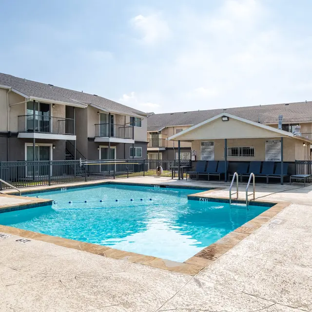 A clear swimming pool surrounded by a concrete deck and residential buildings in the background. Sunlight illuminates the area, and there is a clubhouse near the pool.