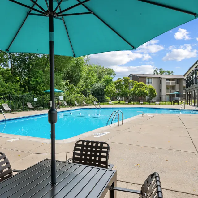 A swimming pool area featuring a clear blue pool surrounded by cement. There are lounge chairs on one side and a dining table with chairs shaded by a turquoise umbrella in the foreground.