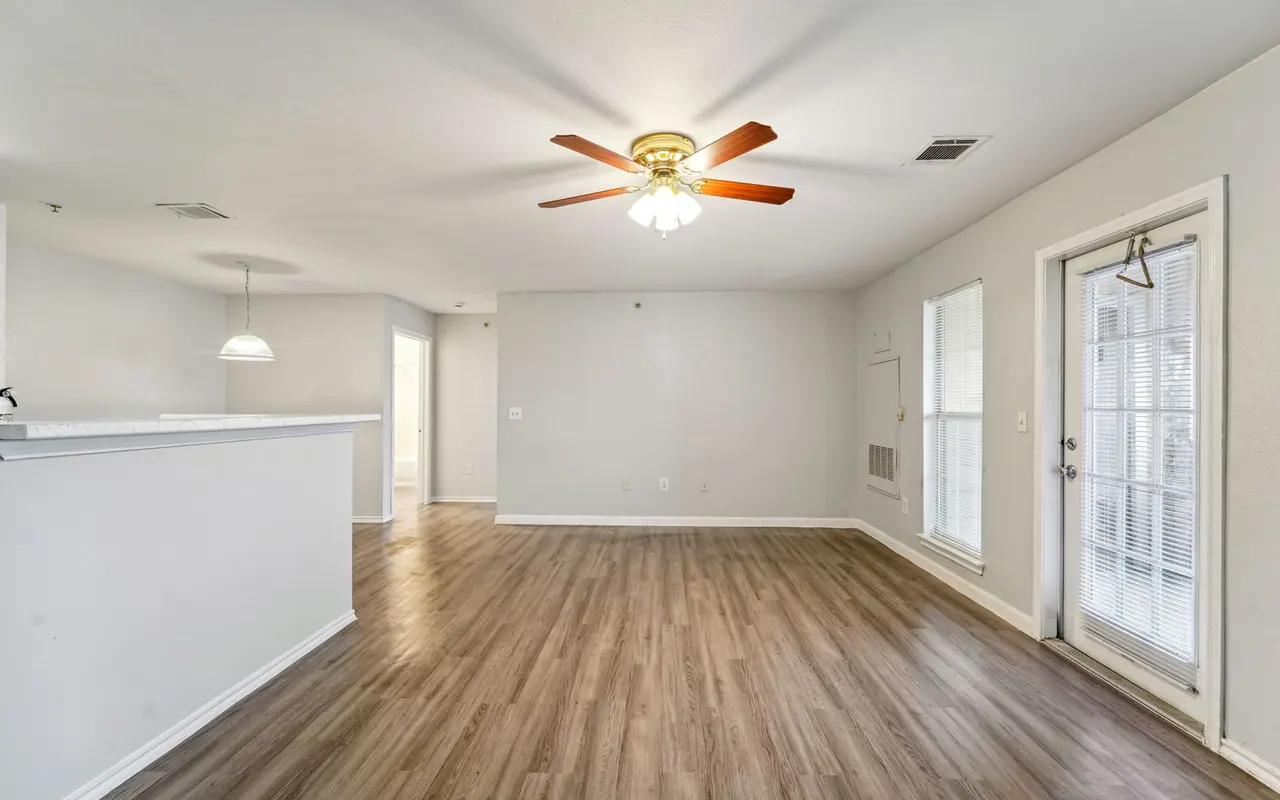 A spacious living room with wooden flooring, a ceiling fan, and a large window with a door leading outside. A partially visible kitchen area is to the left.