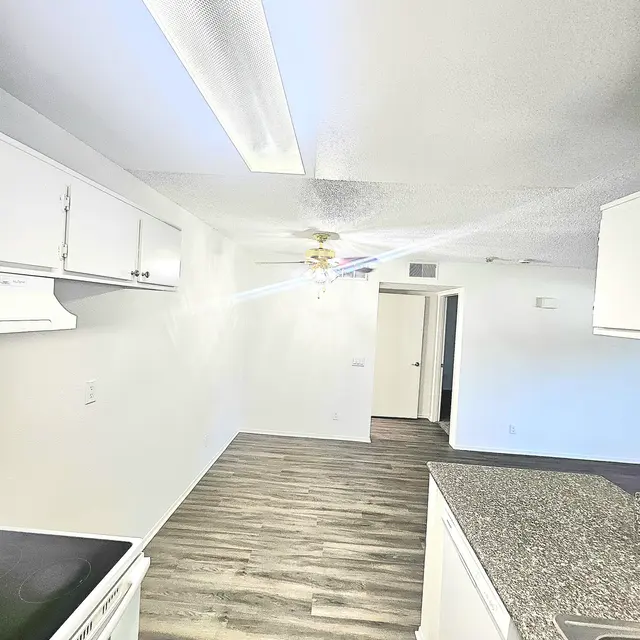 A view of a modern kitchen with white cabinets and a granite countertop. The kitchen features a ceiling fan and an open layout leading to another room.