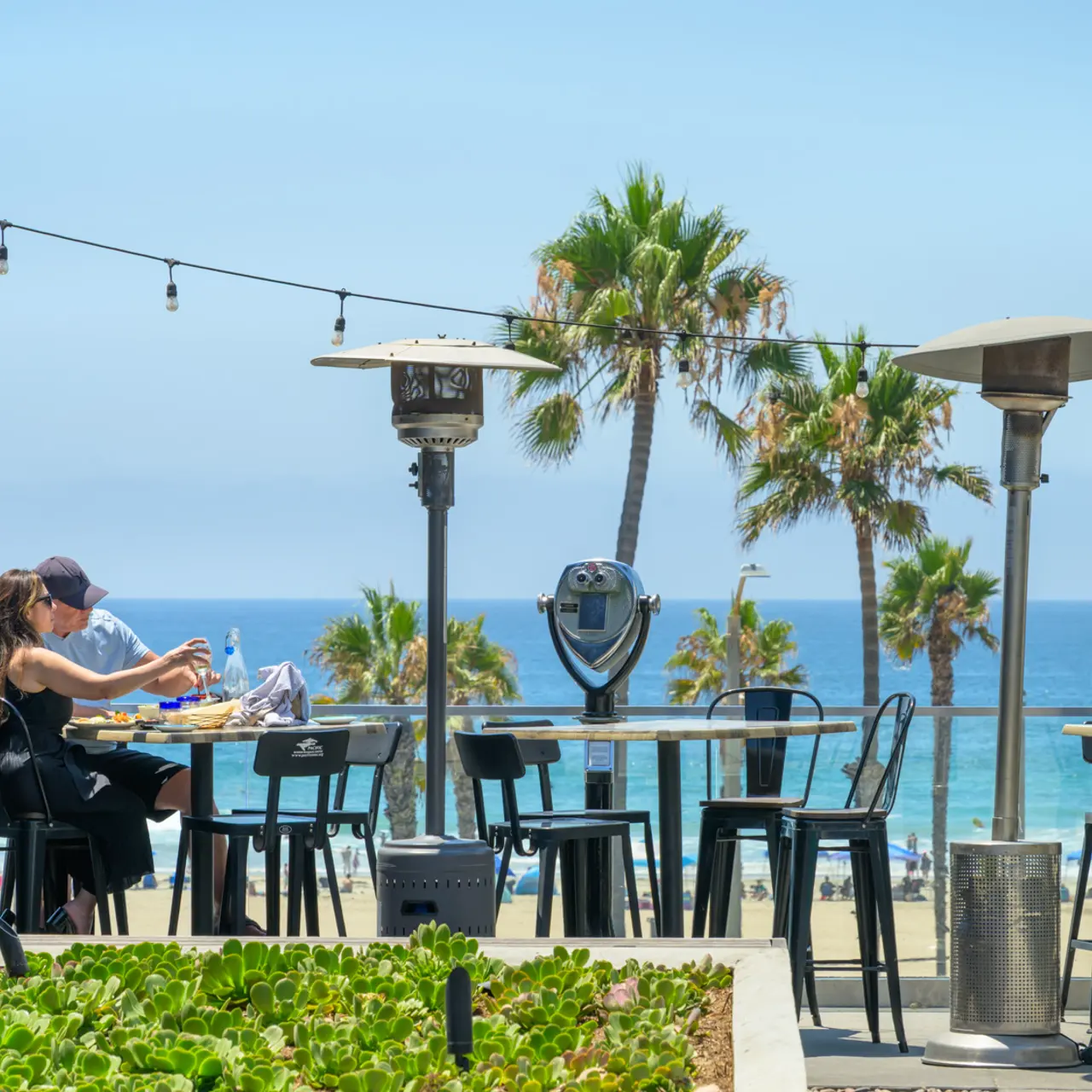 A couple dining outdoors at a beachfront restaurant with palm trees and the ocean in the background.
