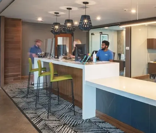 A modern reception area featuring a counter with staff members, bar stools, and decorative lighting fixtures. The space is designed with a blend of wood and contemporary materials.