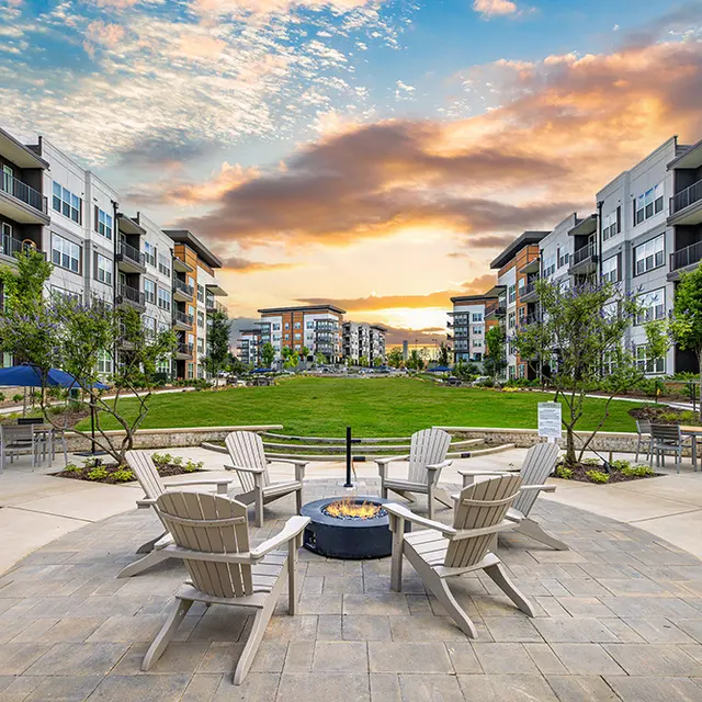 A modern apartment complex courtyard featuring a circular seating area with Adirondack chairs, a fire pit in the center, and landscaped gardens. The background shows several multi-story residential buildings under a colorful sky at sunset.