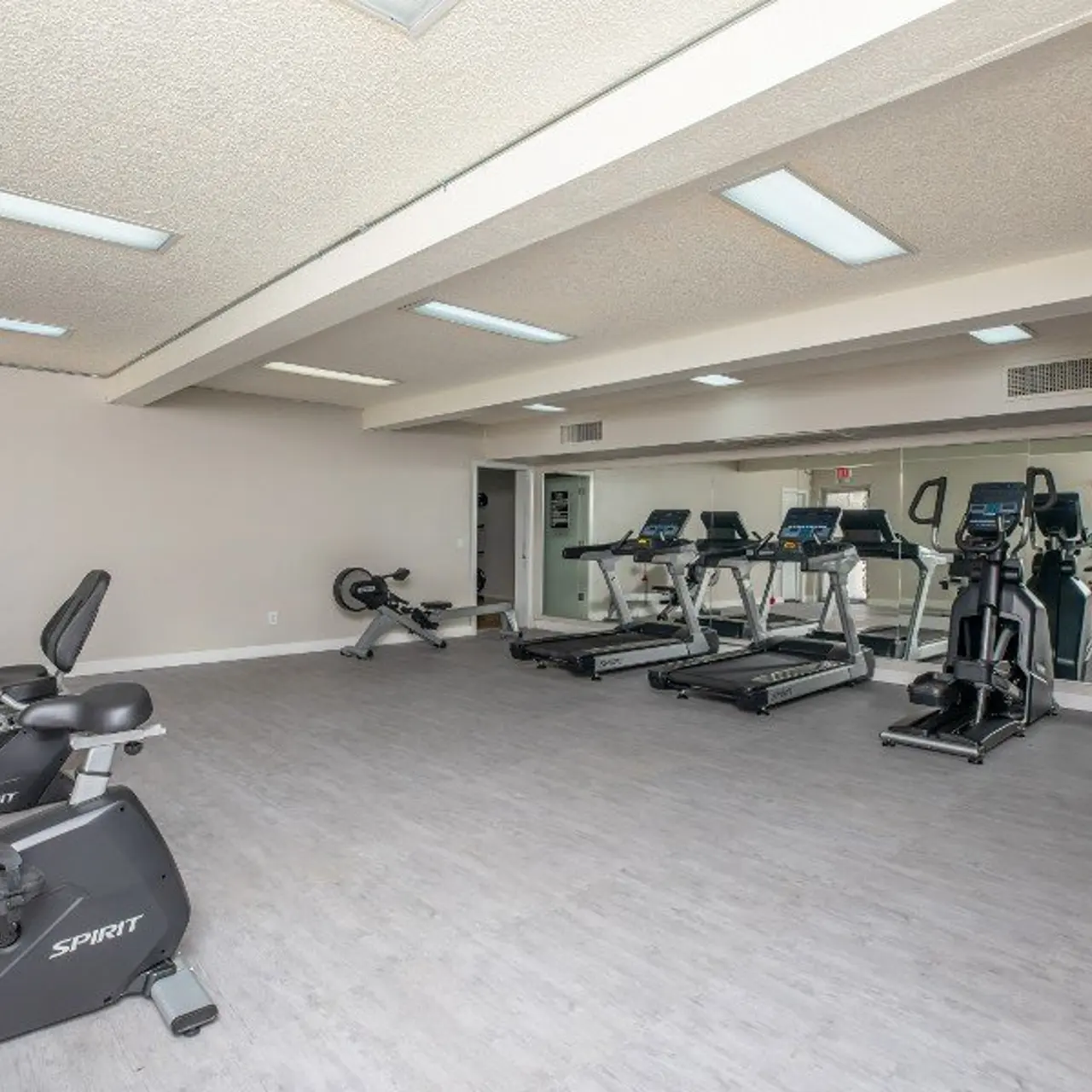 A spacious gym interior featuring exercise equipment including stationary bikes, treadmills, and elliptical machines. The room has a light color scheme with gray flooring and mirrors on one wall.
