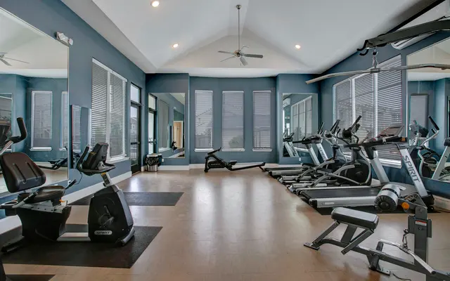 A well-lit, spacious gym featuring various exercise equipment including stationary bikes, treadmills, and weight machines. The decor features blue walls, large windows with shades, and multiple mirrors reflecting the room's setup.