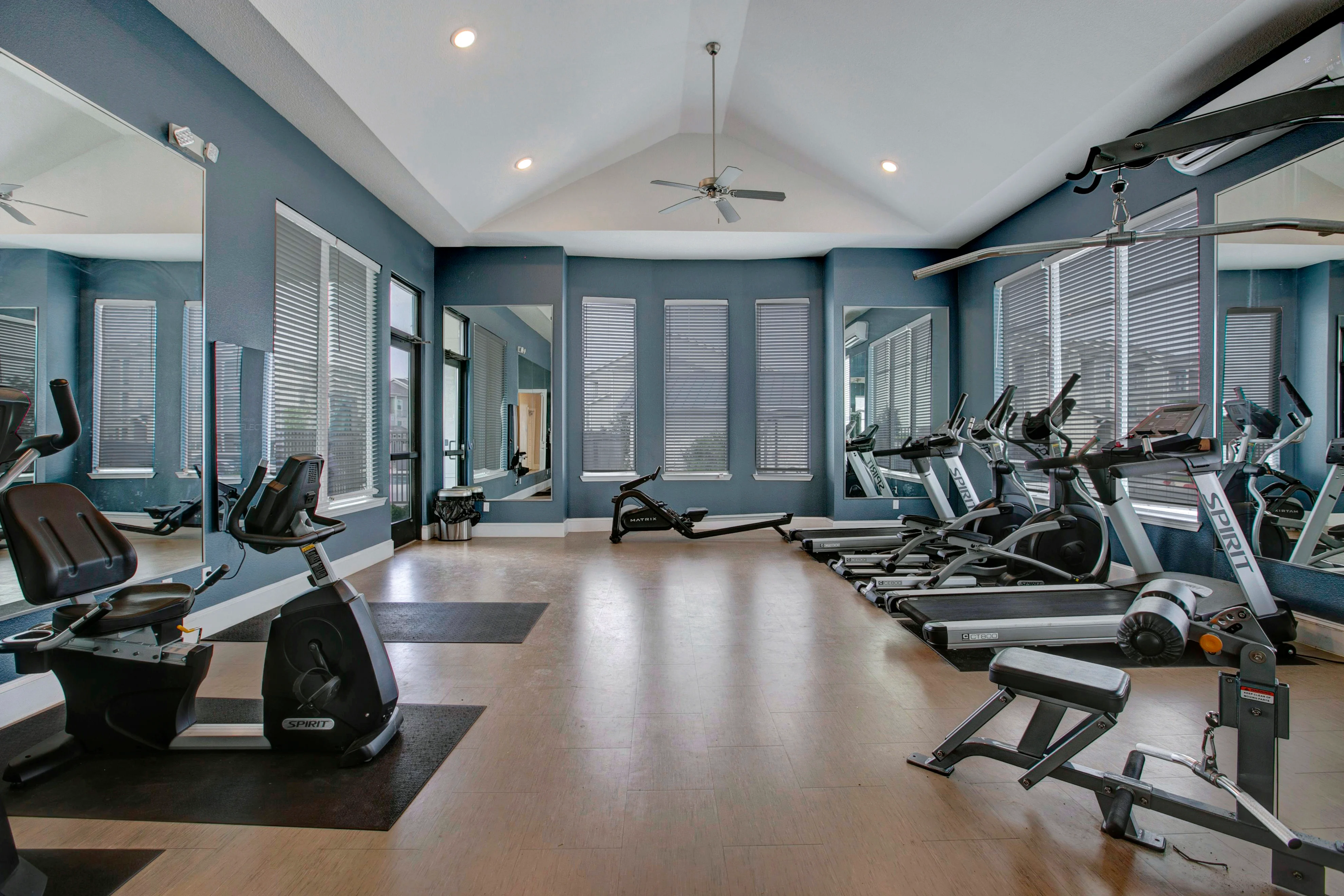 A well-lit, spacious gym featuring various exercise equipment including stationary bikes, treadmills, and weight machines. The decor features blue walls, large windows with shades, and multiple mirrors reflecting the room's setup.