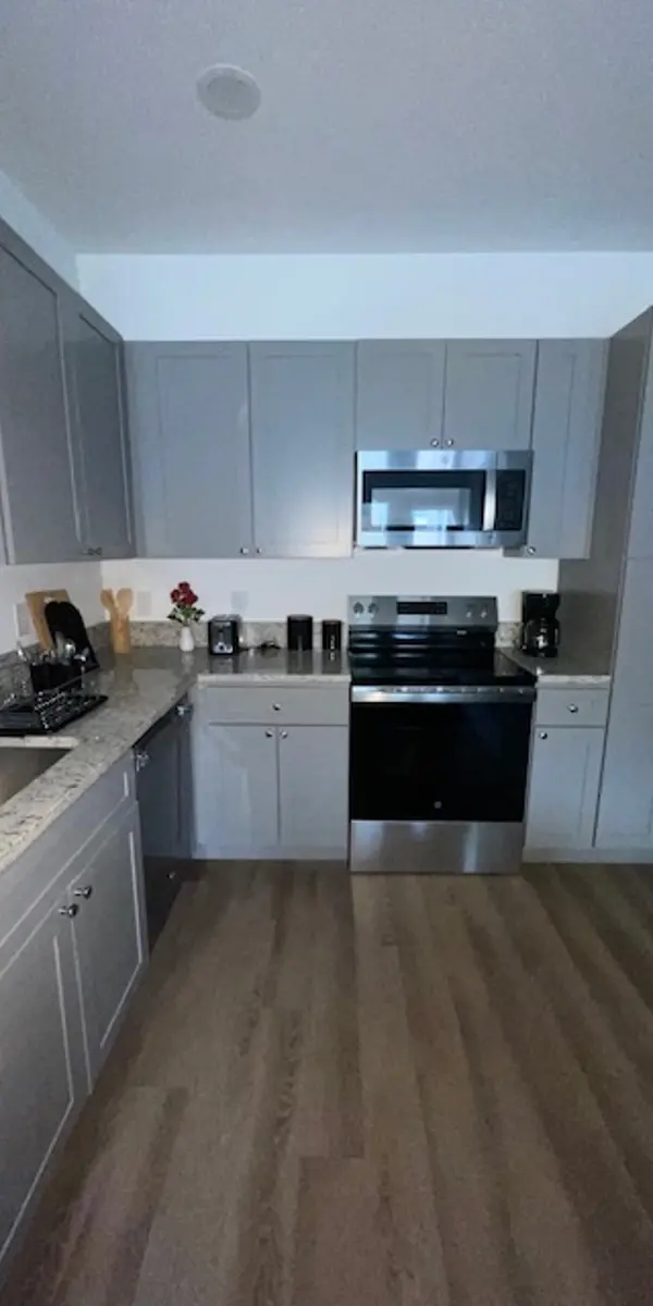 A modern kitchen featuring gray cabinets, stainless steel appliances, and a granite countertop. The kitchen layout includes a sink, stove, and microwave, with wood flooring throughout.